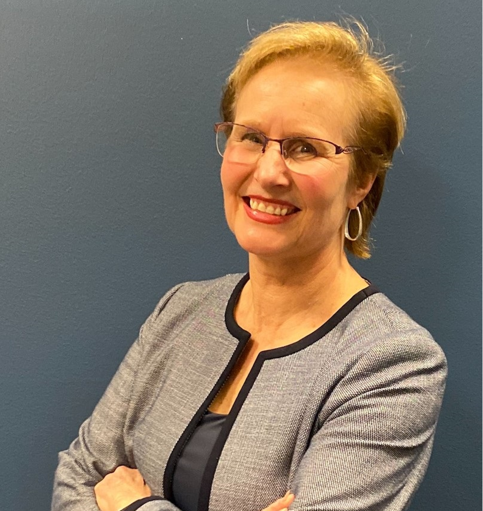 Carolyn Overmyer is posing for a portrait photo. She is wearing a grey blazer and glasses.
