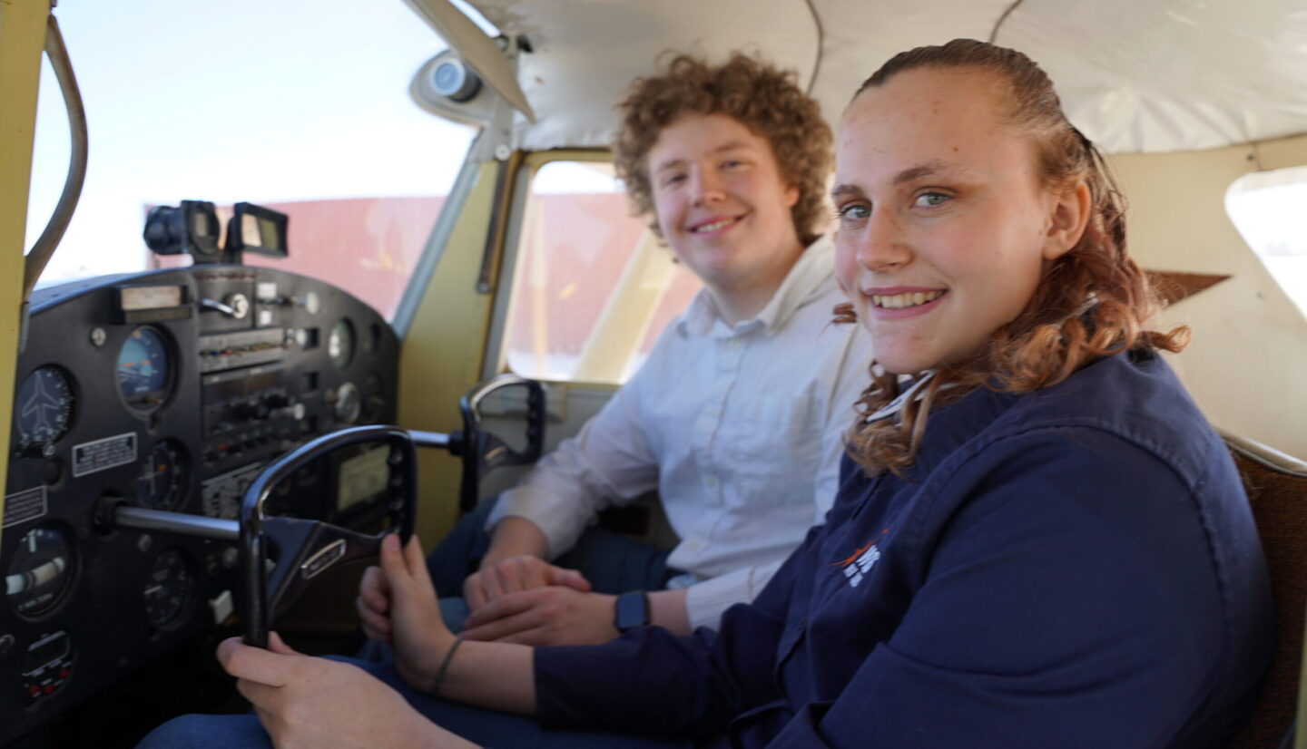 Two people are sitting inside the cockpit of a plane and smiling.