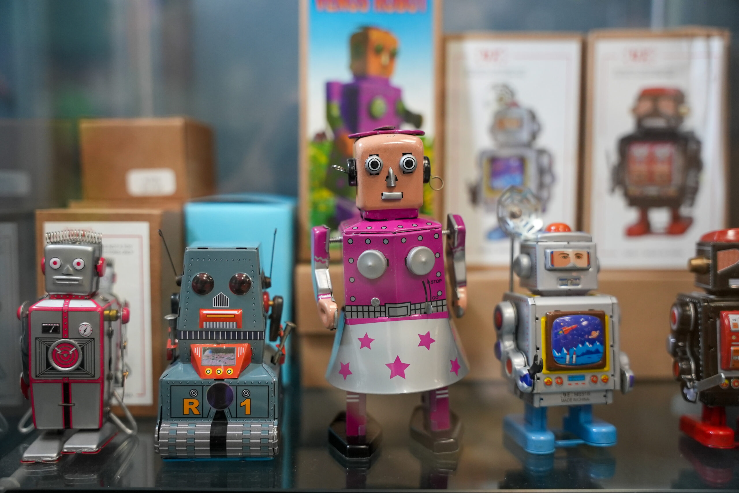 Four square wind-up robots sits on a shelf in front of their boxes.