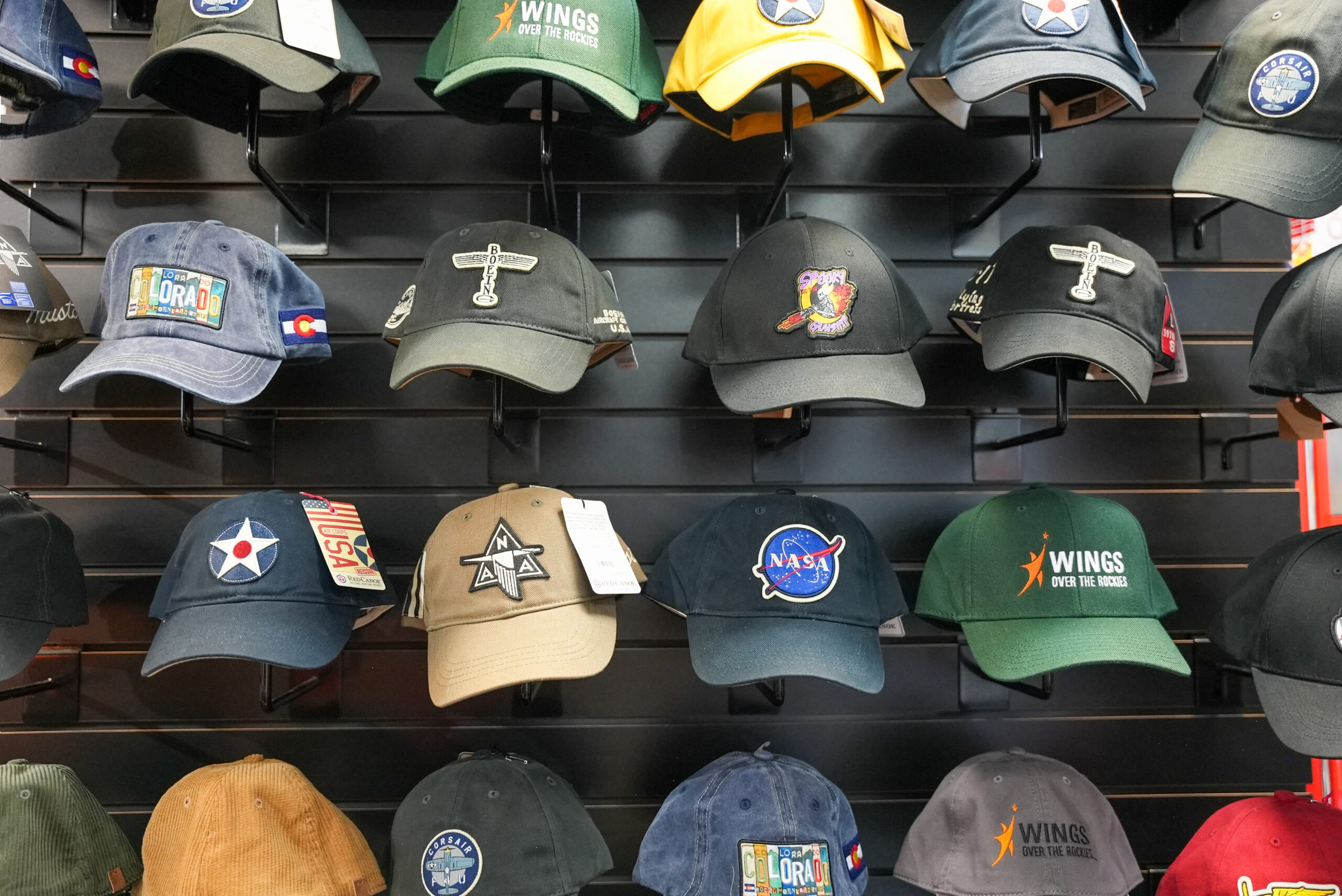 Four rows of various aviation and space related baseball caps hang on a wall.