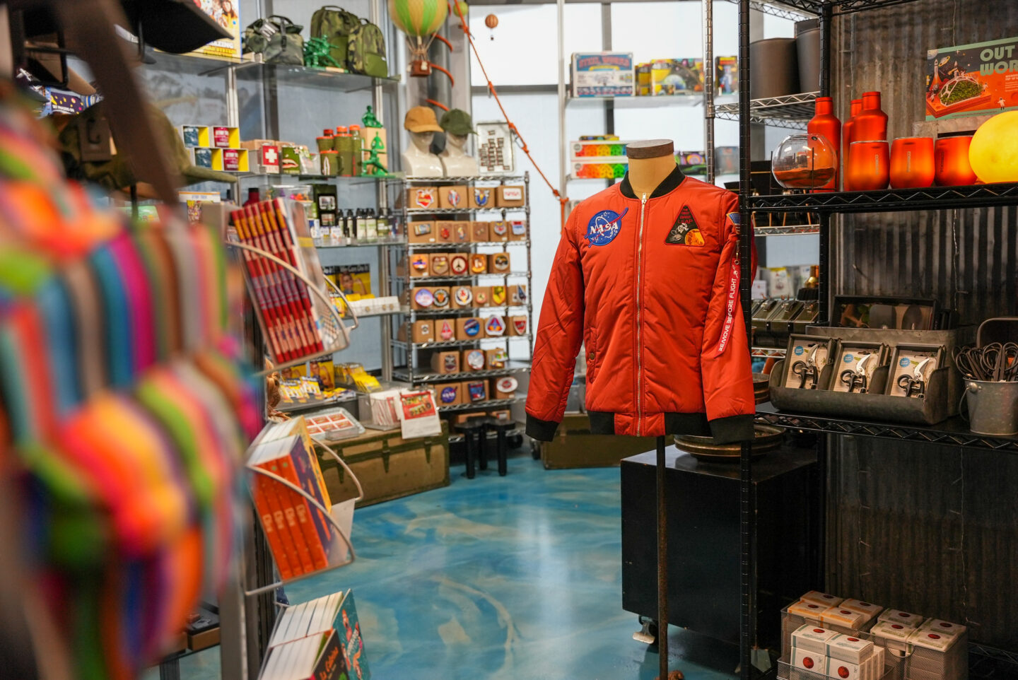 A bright orange bomber jacket with a NASA insignia is displayed on a headless mannequin next to a shelf full of mars related items.