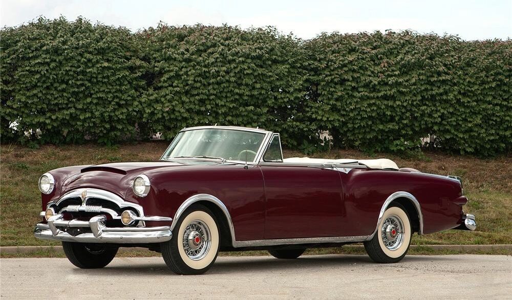 A vintage red car in front of a hedge.