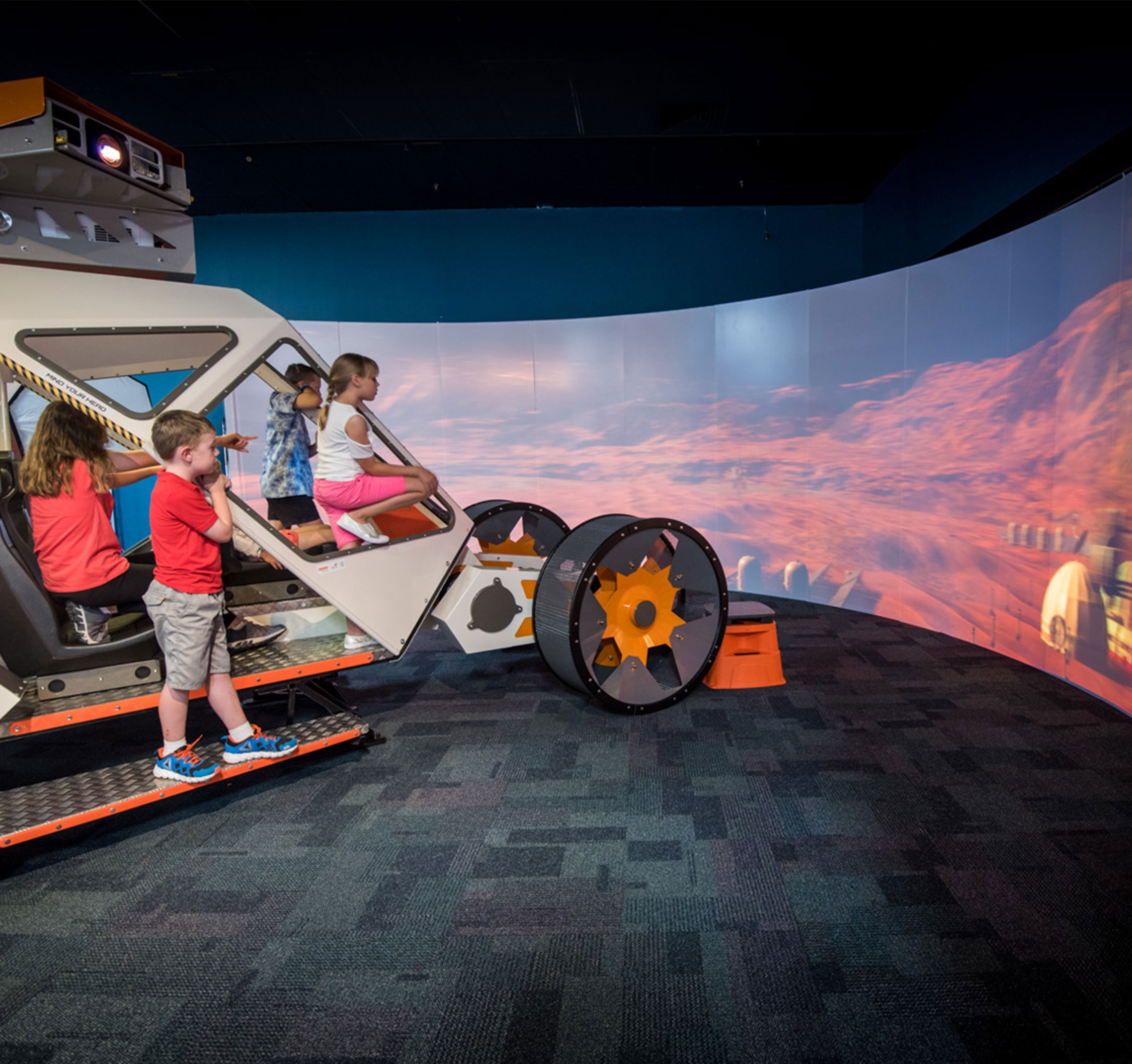 Kids sitting on a Mars Rover replica in front of a Mars-scape